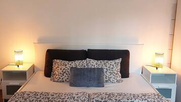 Een net opgemaakt bed met beddengoed en kussens, twee bedlampen en een stadsgezichtschilderij aan de muur.