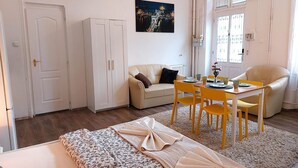 A room with a bed, a sofa, a dining table with chairs, a wardrobe, and a window with blinds. - GoodMo Budapest Smart Rooms (Budapest)