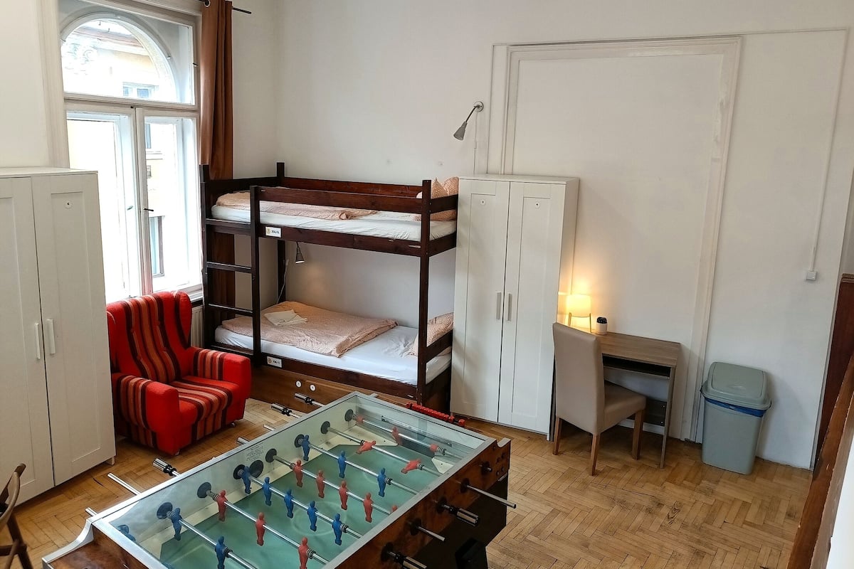 A dormitory room with bunk beds, a foosball table, a desk, and a chair.