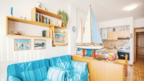 A cozy living room with a blue-striped sofa, wooden shelves with books and decorative items, a sailboat model, and a kitchen area with wooden cabinets and a stove.