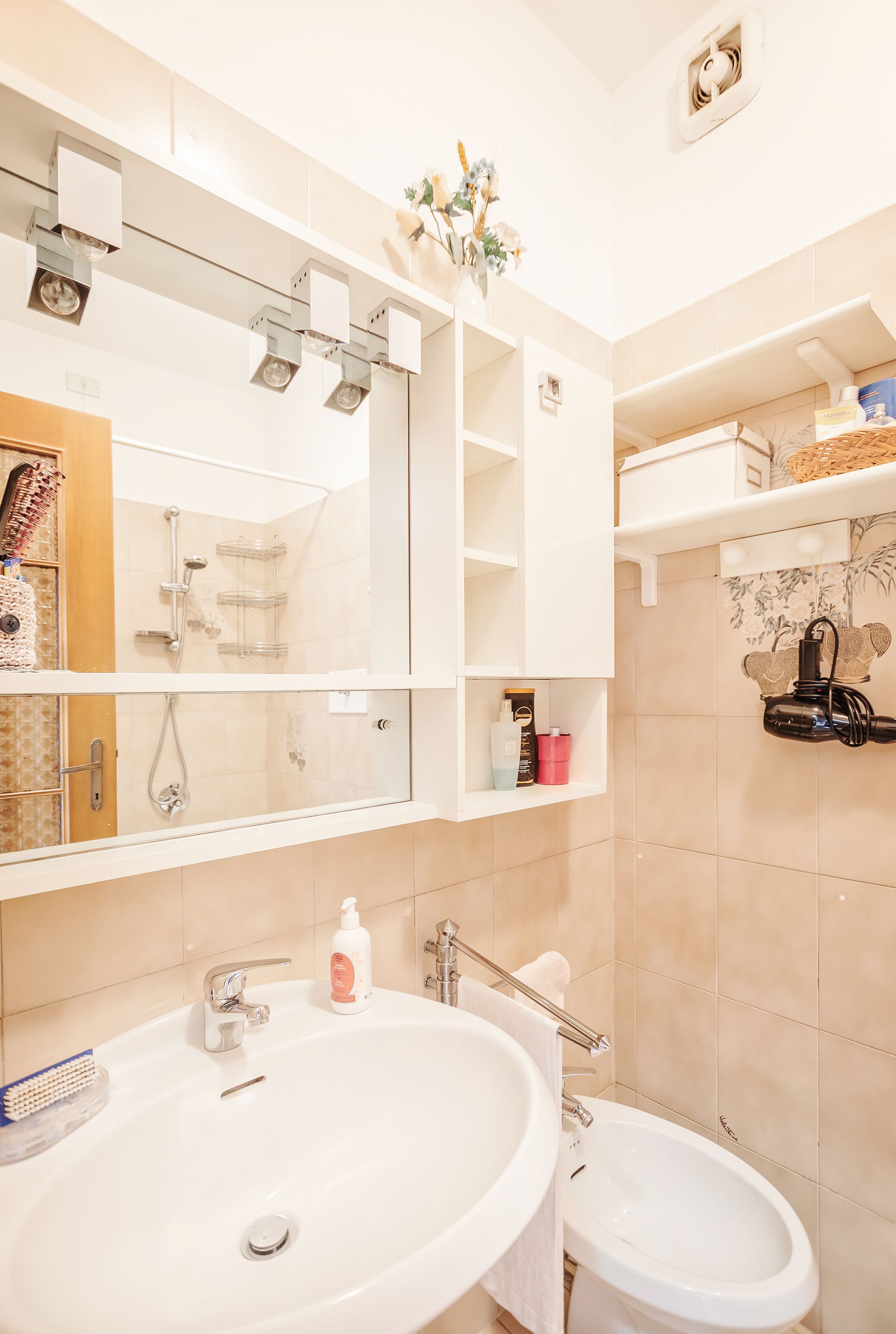 A bathroom with a large mirror, a sink, a bidet, and shelves with toiletries.