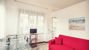 A living room with a red sofa, a glass dining table with chairs, a TV on a stand, and a beach-themed poster on the wall. - Sanremo Central Nest by Wonderful Italy (Sanremo)