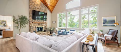 A spacious living room with a stone fireplace, a large sectional sofa, and a high ceiling with wooden beams.