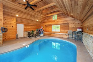 An indoor swimming pool with wooden walls and ceiling, a ceiling fan, and a seating area with chairs and a table.