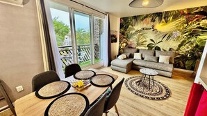 A modern living room with a large sliding glass door leading to a balcony, a grey sofa set, a wooden dining table with woven placemats, and a tropical-themed mural on the wall. - Bikini & Martini au Manganao Pool XXL & Plage (Saint-Francois)