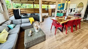 Een ruime woonkamer met een bank, een eettafel met rode stoelen, een keukenhoek en een groot raam met uitzicht op groen.