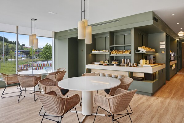 Un comedor moderno con sillas de mimbre y una mesa redonda, una barra con comida y bebidas, y ventanas amplias que ofrecen vistas al exterior.