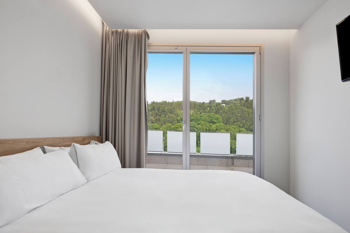 A modern hotel room with a large bed, a view of a forest through a sliding glass door, and a flat-screen TV mounted on the wall.