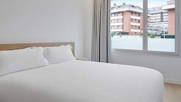 Una cama con sábanas blancas en una habitación con cabecera de madera, una mesita de noche con lámpara y una ventana con vistas a edificios.