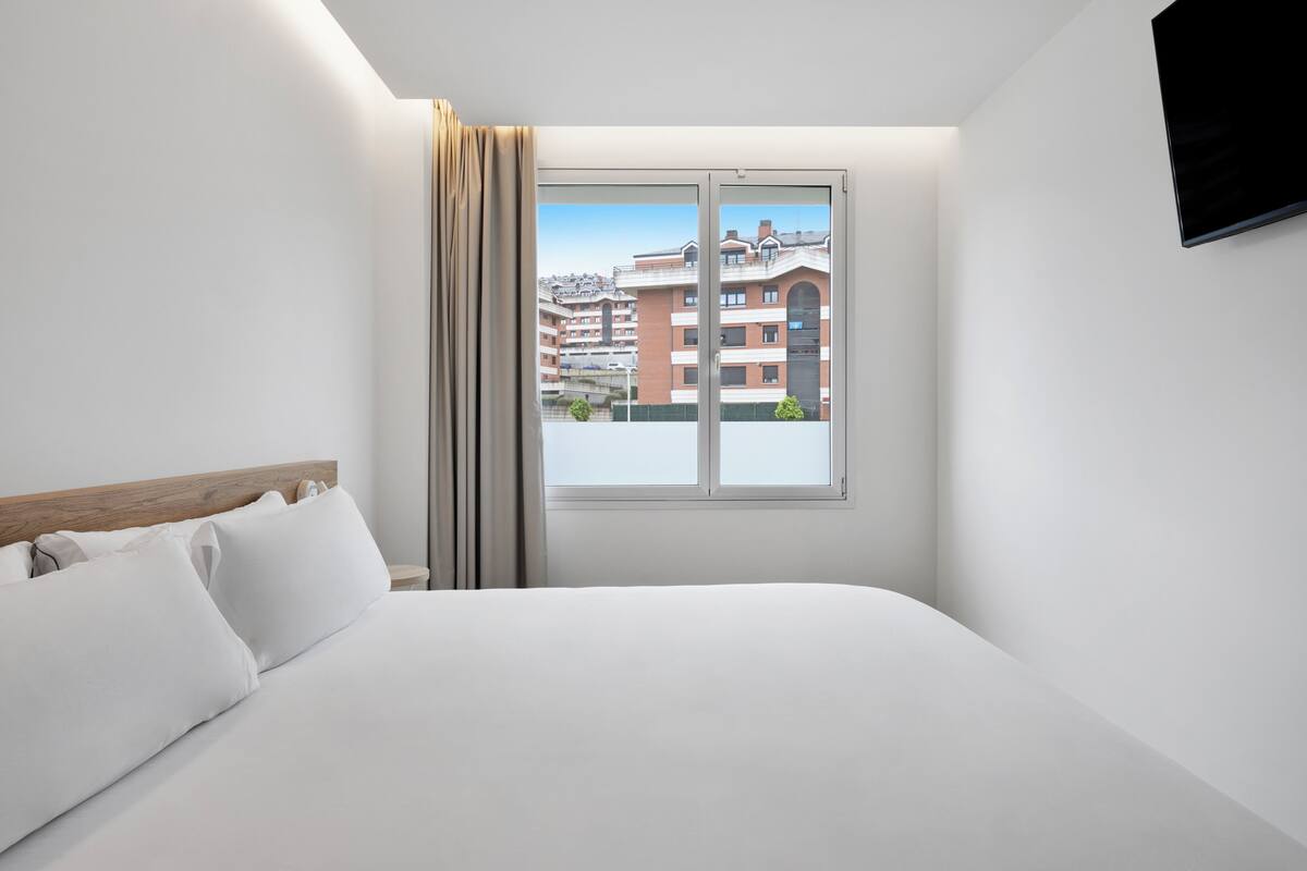 A hotel room with a large bed, a window with curtains, and a view of buildings outside.