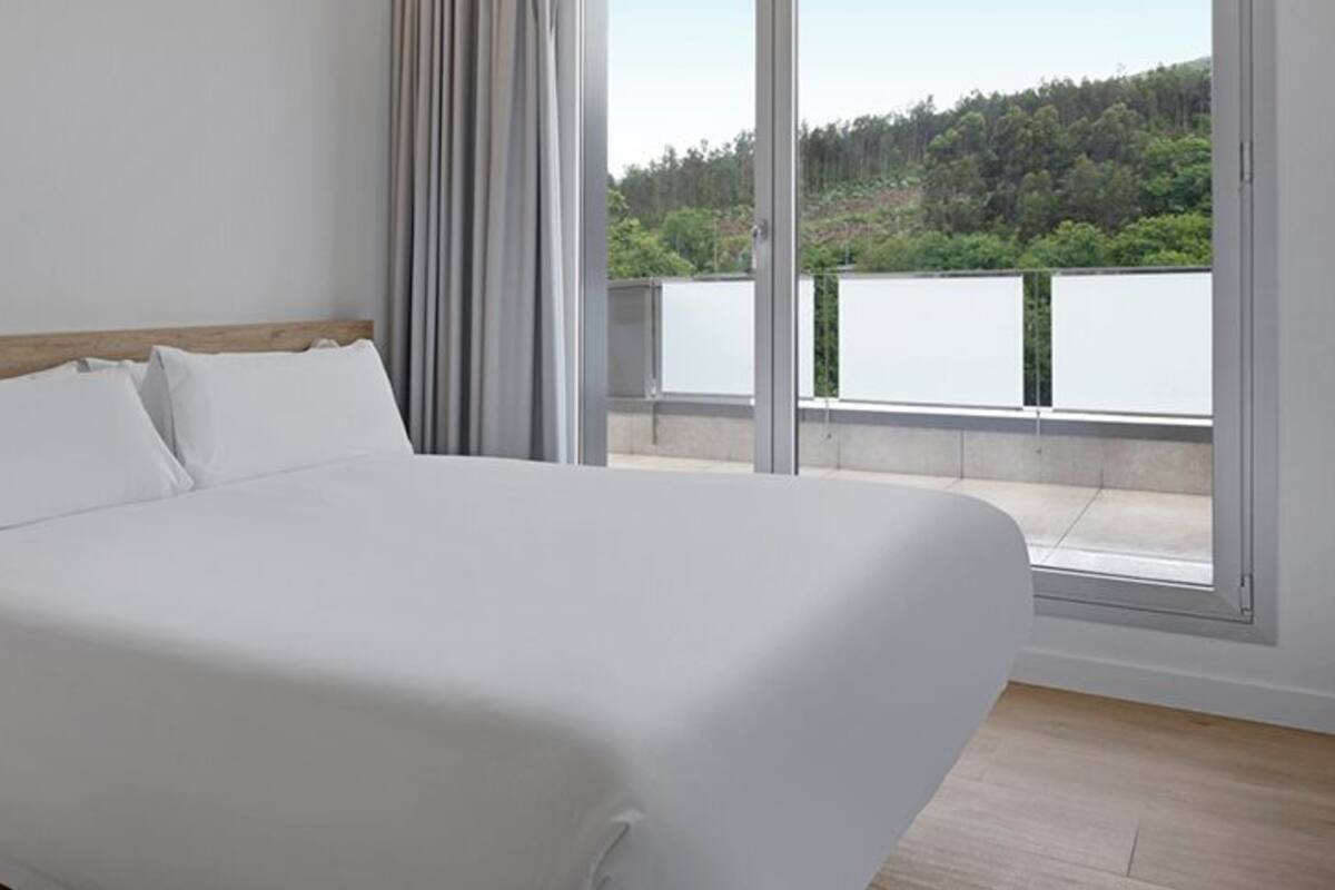 A modern hotel room with a large bed, a bedside table, and a view of a lake through large windows.