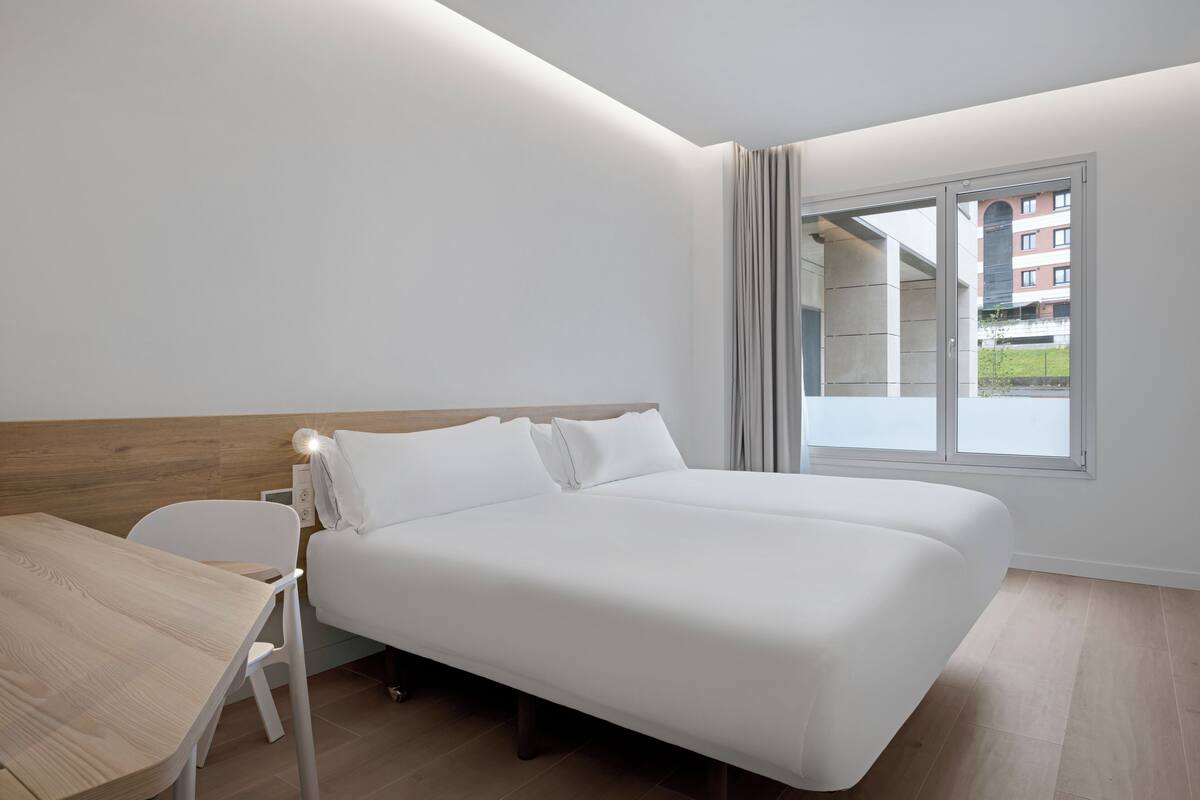 A modern hotel room with a large bed, a wooden desk, and a window with a view of buildings.