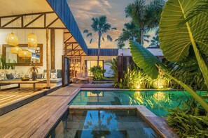 Un área de piscina moderna al aire libre con cubierta de madera, sillas de descanso y plantas tropicales.