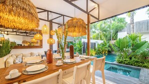 Un comedor con una mesa de madera preparada para cuatro personas, rodeada de sillas blancas, con una piscina y exuberante follaje visibles a través de las paredes de vidrio.