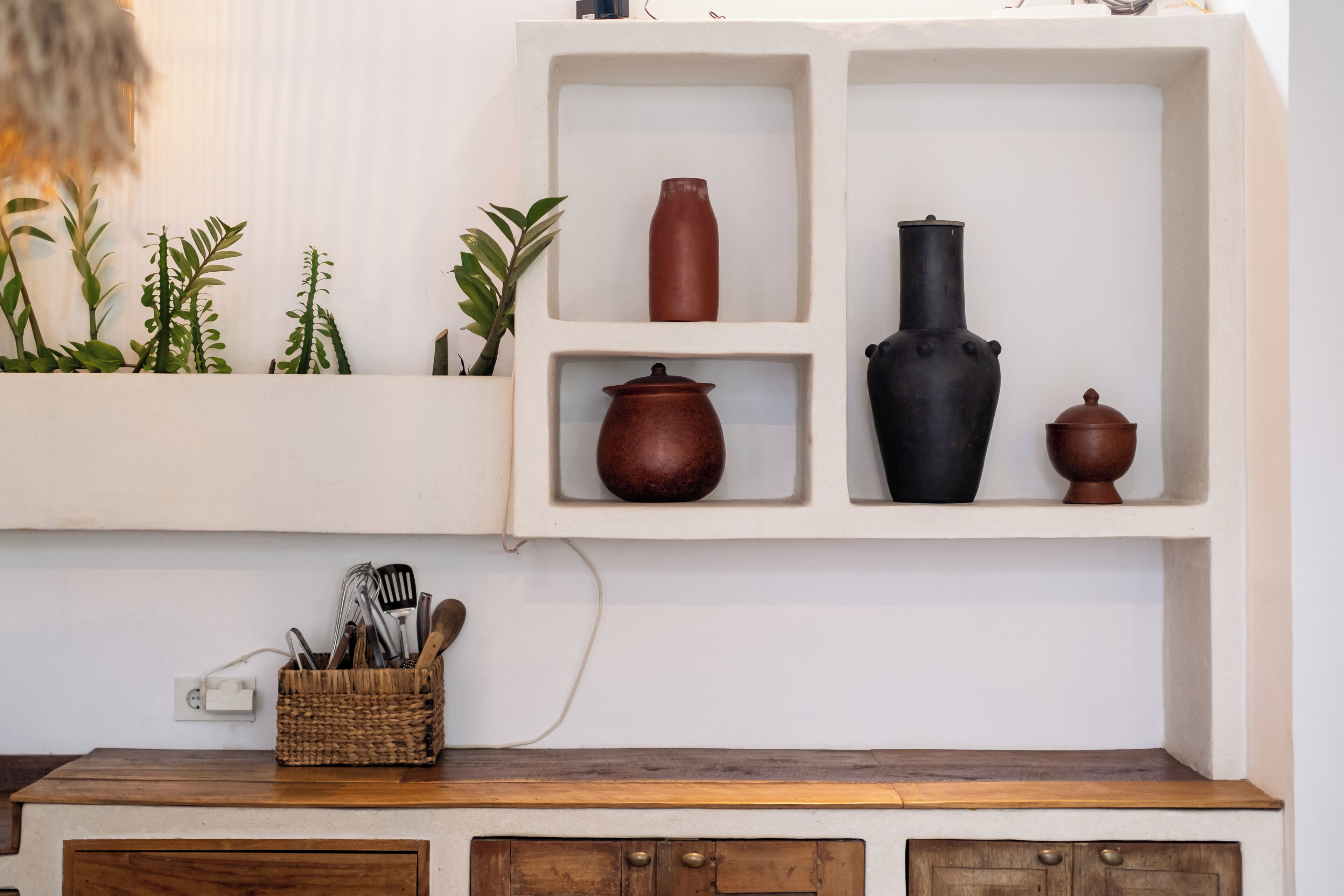 Une cuisine avec un plan de travail en bois, une étagère blanche sur laquelle sont posés trois objets en céramique, et un panier rempli d’ustensiles.