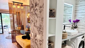 A modern room with a wooden floor, a sofa with a yellow cushion, a decorative wall with a tropical pattern, and a washing machine.