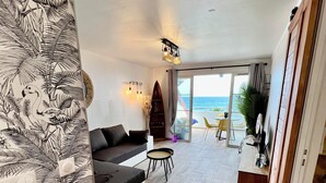 En moderne stue med sofa, et lille bord og en surfbræt. Stort vindue med udsigt over havet.