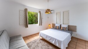 A bedroom with a bed, a nightstand, a lamp, a window with curtains, and a view of greenery outside. - Villa Chill & Ride Le Helleux Magnifique Piscine (Sainte-Anne)