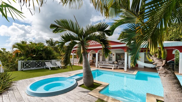 A swimming pool with a wooden deck, palm trees, and a red building with white columns in the background. - Villa Chill & Ride Le Helleux Magnifique Piscine (Sainte-Anne)