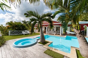 A swimming pool with a wooden deck, palm trees, and a red building with white columns in the background. - Villa Chill & Ride Le Helleux Magnifique Piscine (Sainte-Anne)