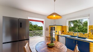 A kitchen with a stainless steel refrigerator, a wooden dining table with chairs, a hanging light fixture, and a window with a view of greenery. - Villa Chill & Ride Le Helleux Magnifique Piscine (Sainte-Anne)