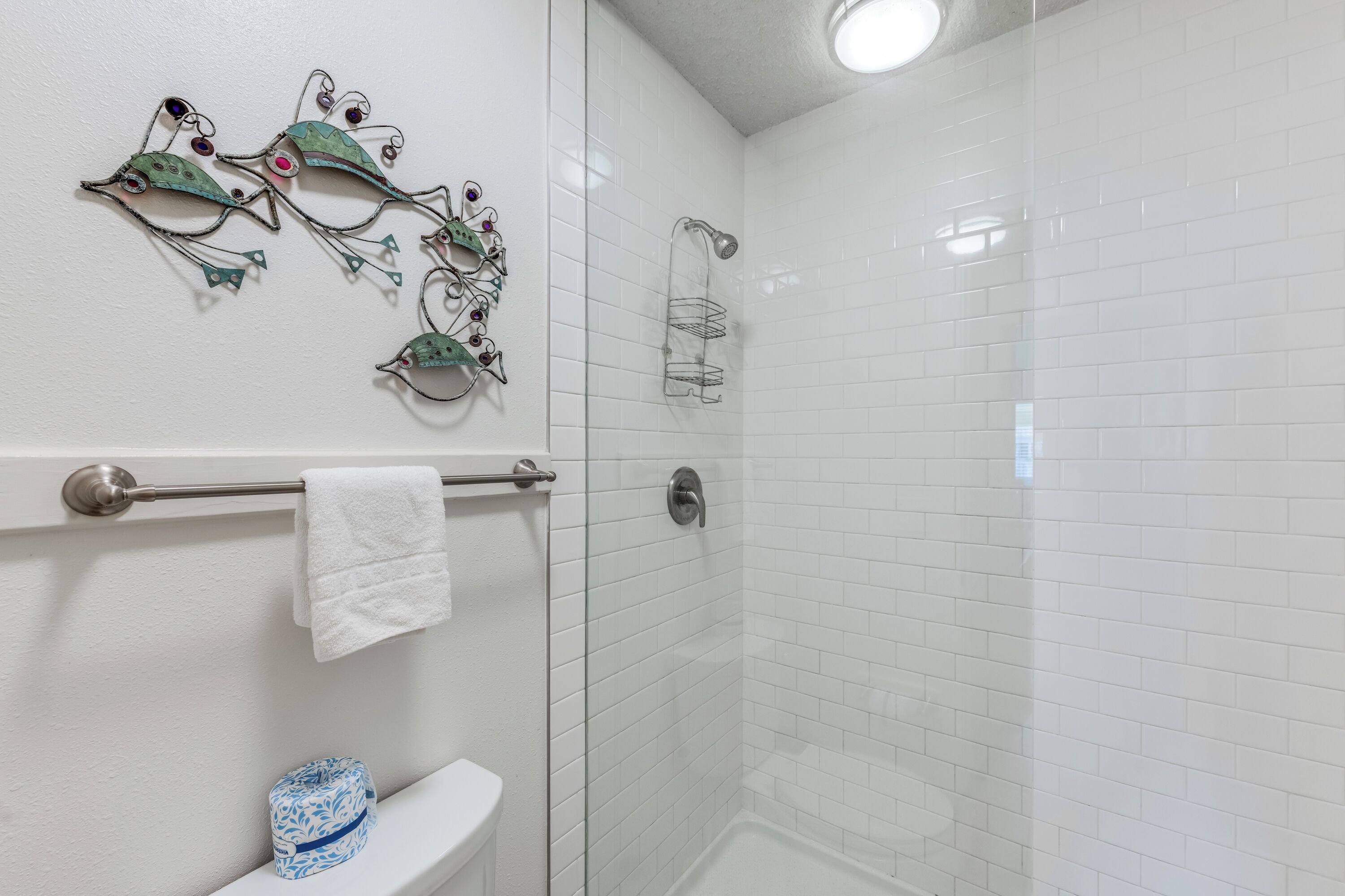 Un bagno con pareti piastrellate in bianco, una doccia con porta in vetro, un porta asciugamani con un asciugamano bianco e un elemento decorativo a parete.