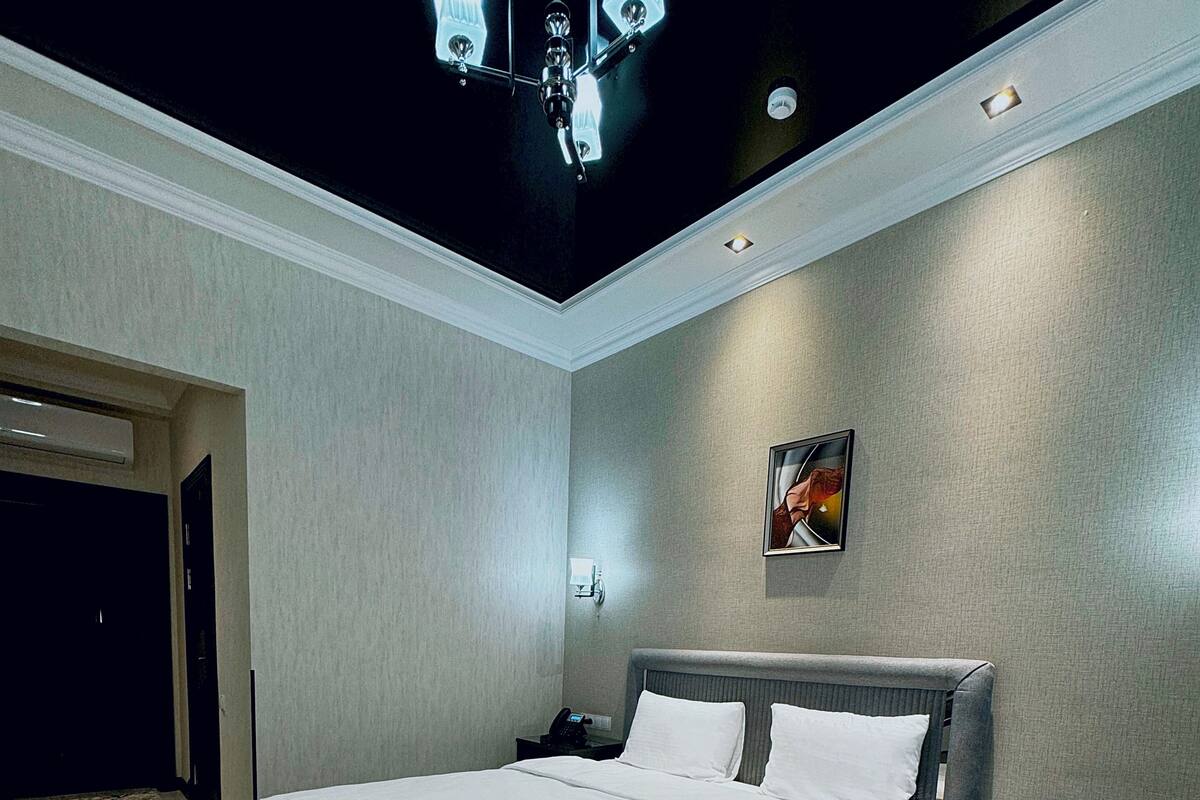 A modern hotel room with a large bed, a nightstand, a desk, and a ceiling light fixture.