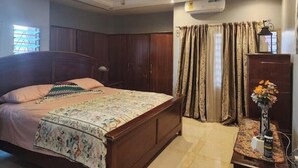 Une chambre à coucher comprenant un lit, un ventilateur de plafond, un appareil de climatisation, des rideaux, une table de chevet avec une lampe et un vase avec des fleurs.