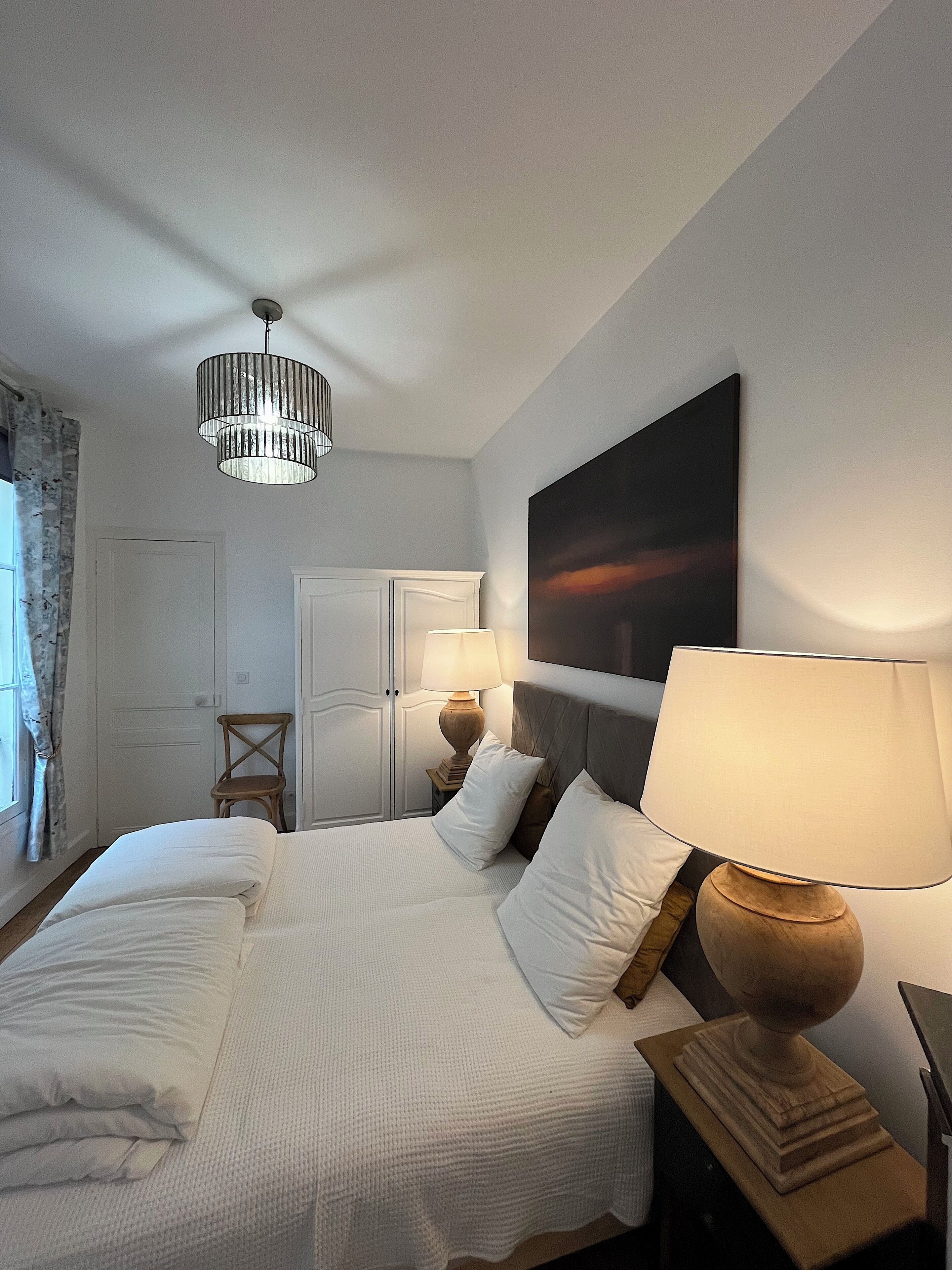 A hotel room with a bed, a nightstand with a lamp, a wooden chair, a wardrobe, and a painting on the wall.