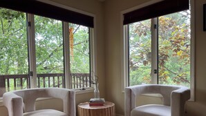 A room with two white armchairs, a small wooden side table with a vase and books, and a view of trees through large windows. - Beautiful family home in the Upper Rattlesnake (Missoula)