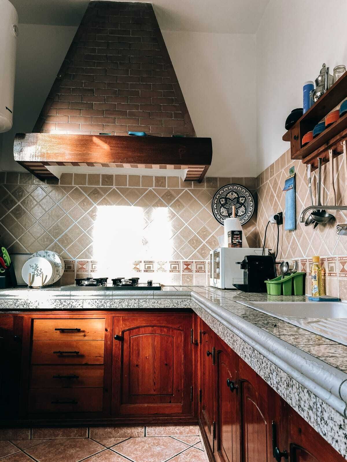 A kitchen with a brick chimney, tiled backsplash, wooden cabinets, and a countertop with various kitchen items.