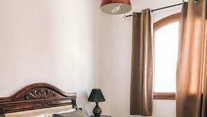 A neatly made bed with a wooden headboard, a bedside lamp, and a window with curtains. - Amber apartment  (Essaouira)
