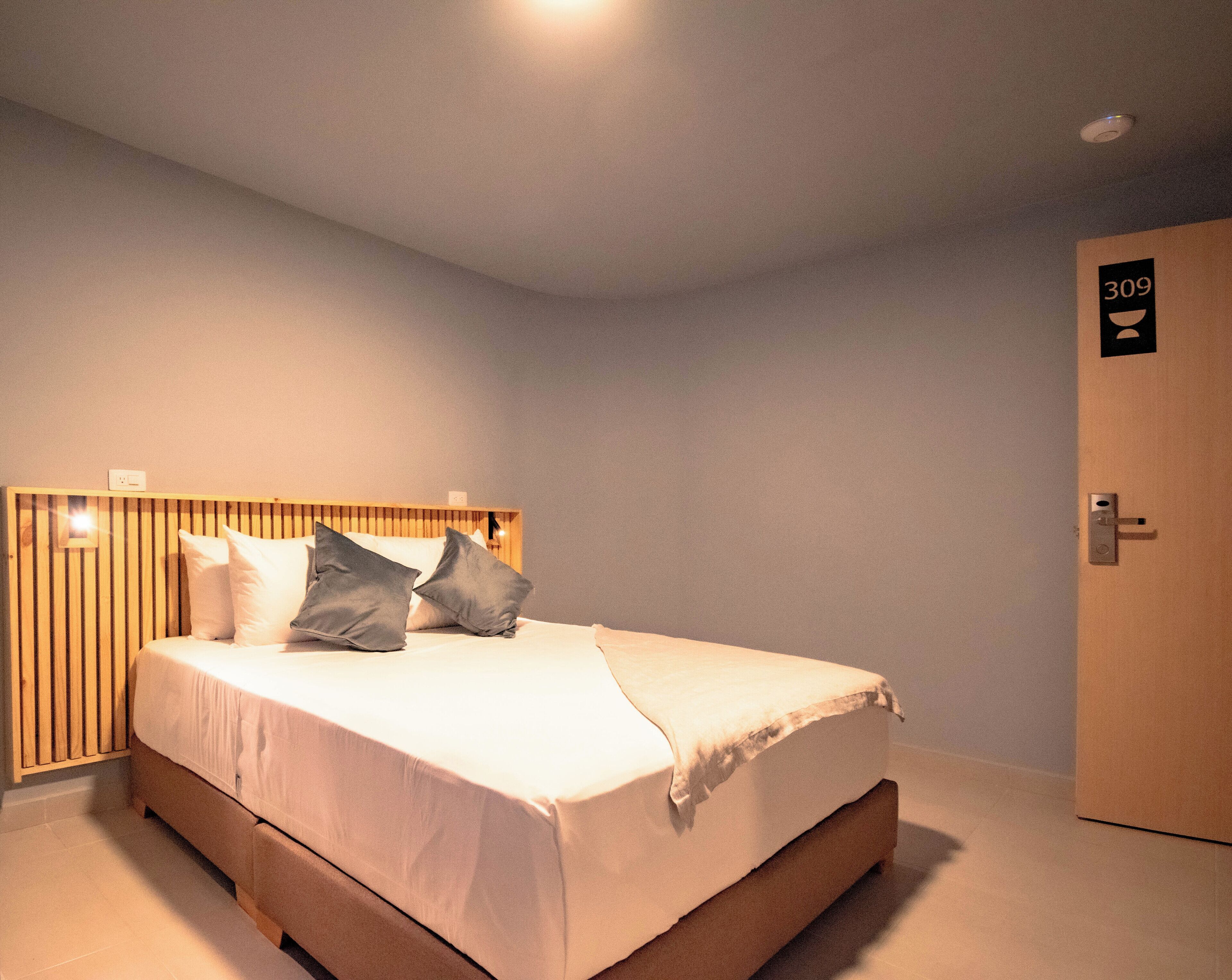 A neatly made bed with a wooden headboard, a door with a number sign, and a light switch on the wall.
