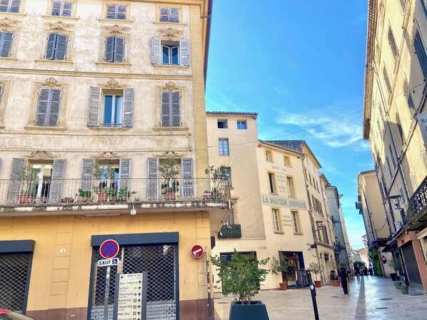 A narrow street with buildings featuring balconies with potted plants, a sign for 'Hotel de Ville,' and a 'No Parking' sign.