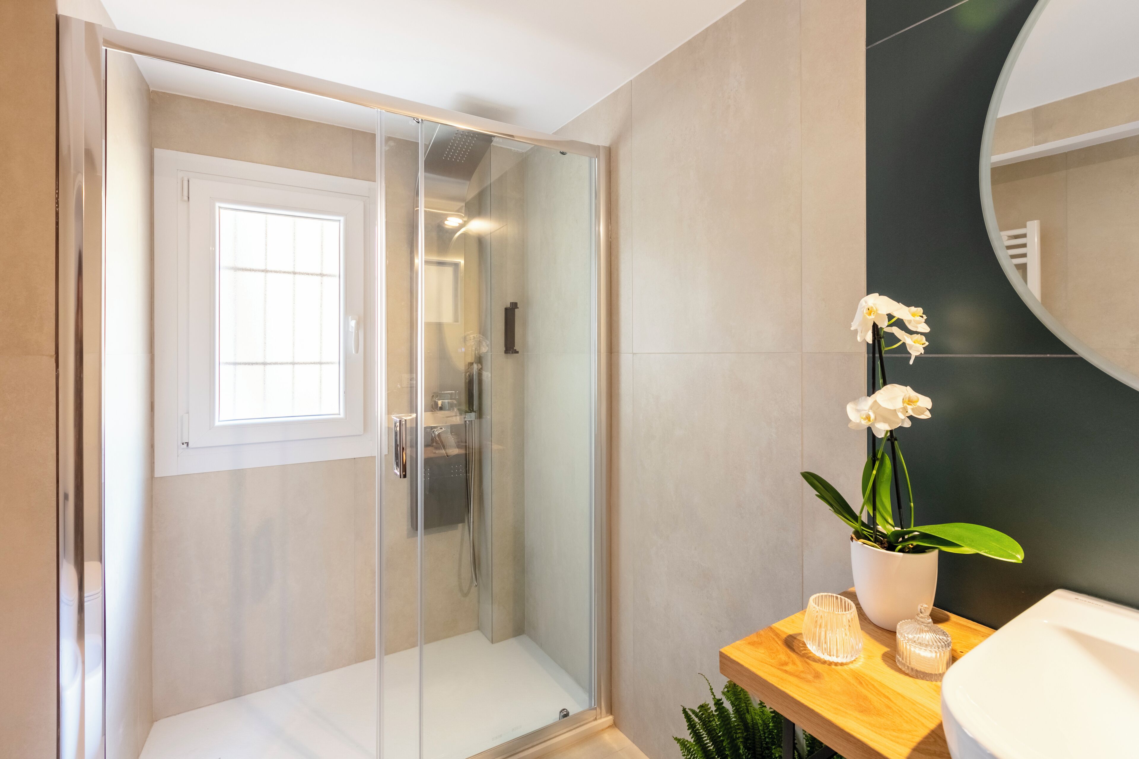 Une salle de bain moderne avec une douche à parois de verre, un lavabo blanc et une orchidée en pot posée sur une étagère en bois.