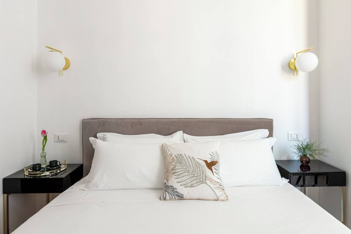 A neatly made bed with white linens and two pillows, one with a leaf pattern. There are two bedside tables, one with a vase of flowers and a small plant, and the other with a decorative tray.