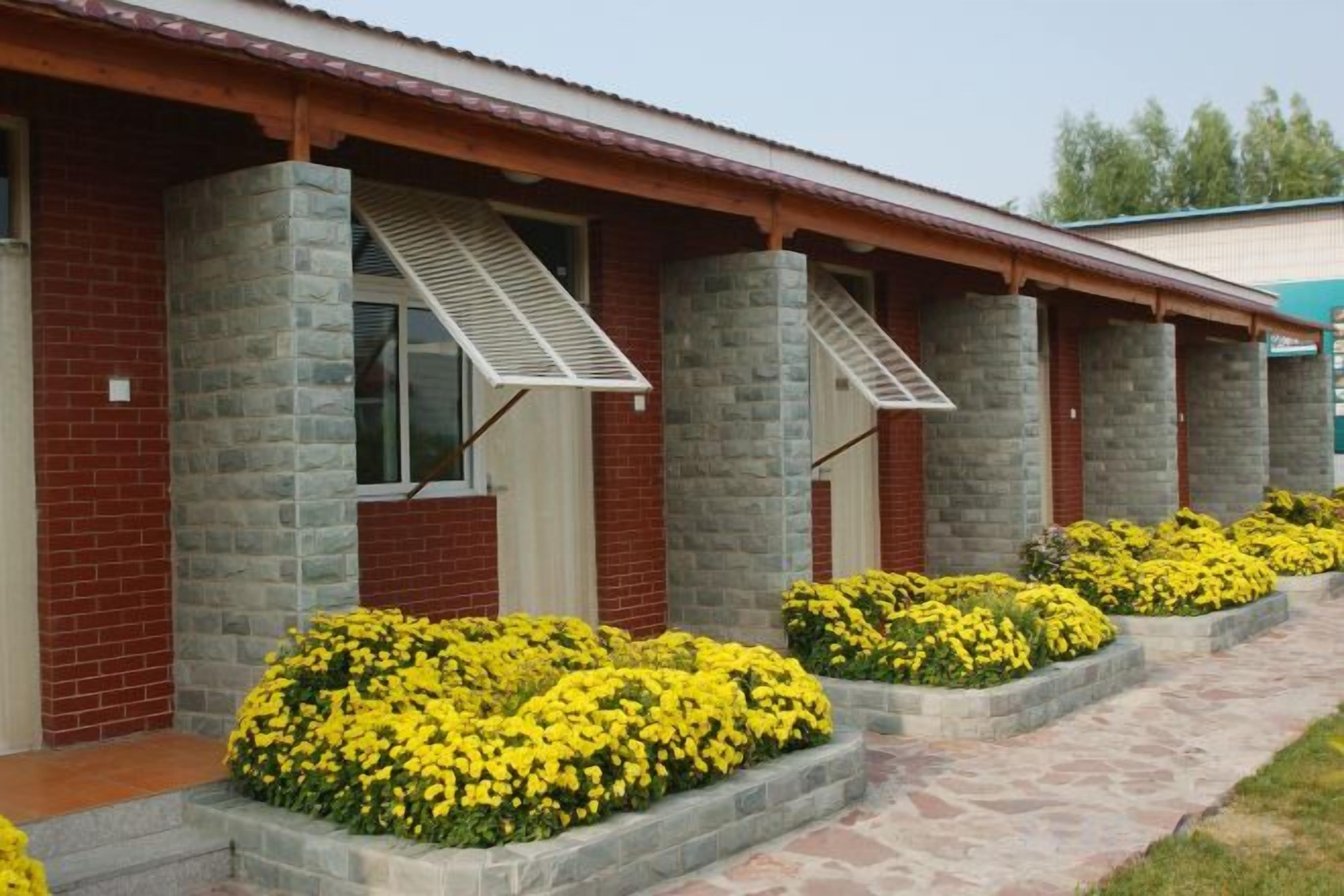 Una hilera de habitaciones con pilares de piedra, ventanas con toldos y macizos de flores amarillas.