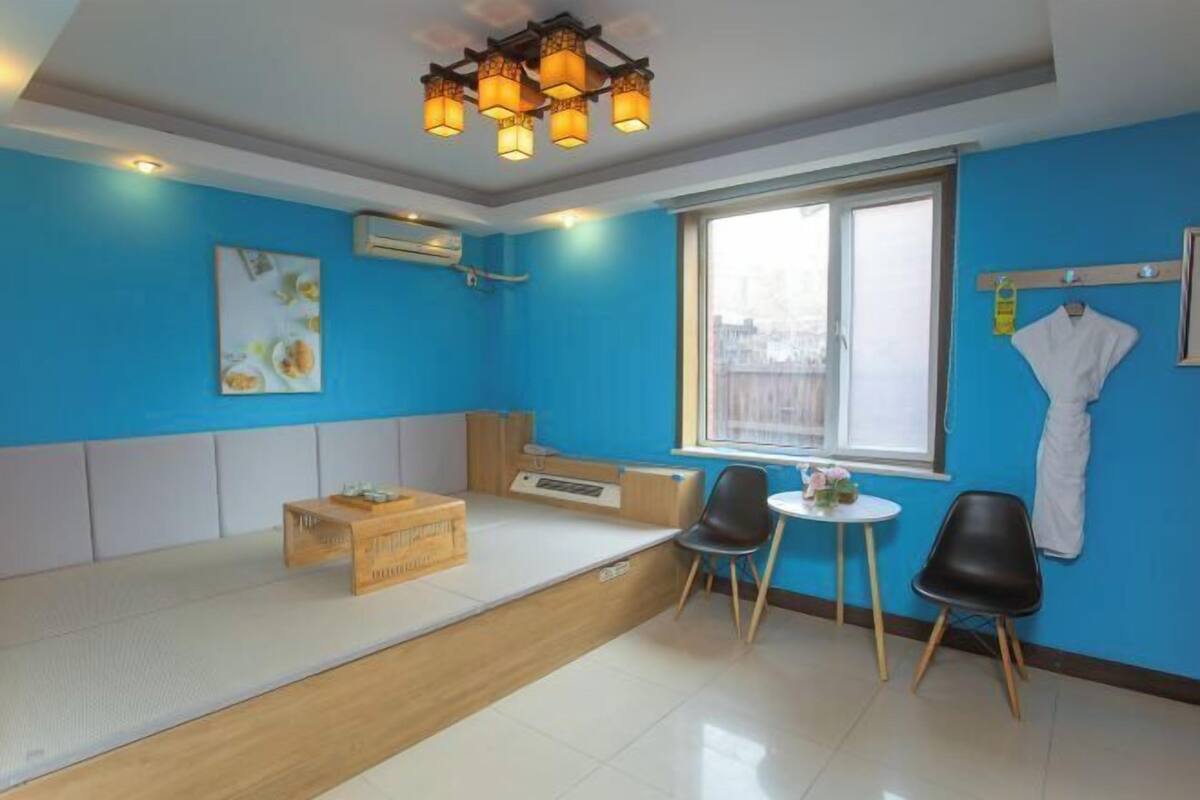 A room with a blue wall, a window, a bed, a small table with chairs, and a ceiling light fixture.