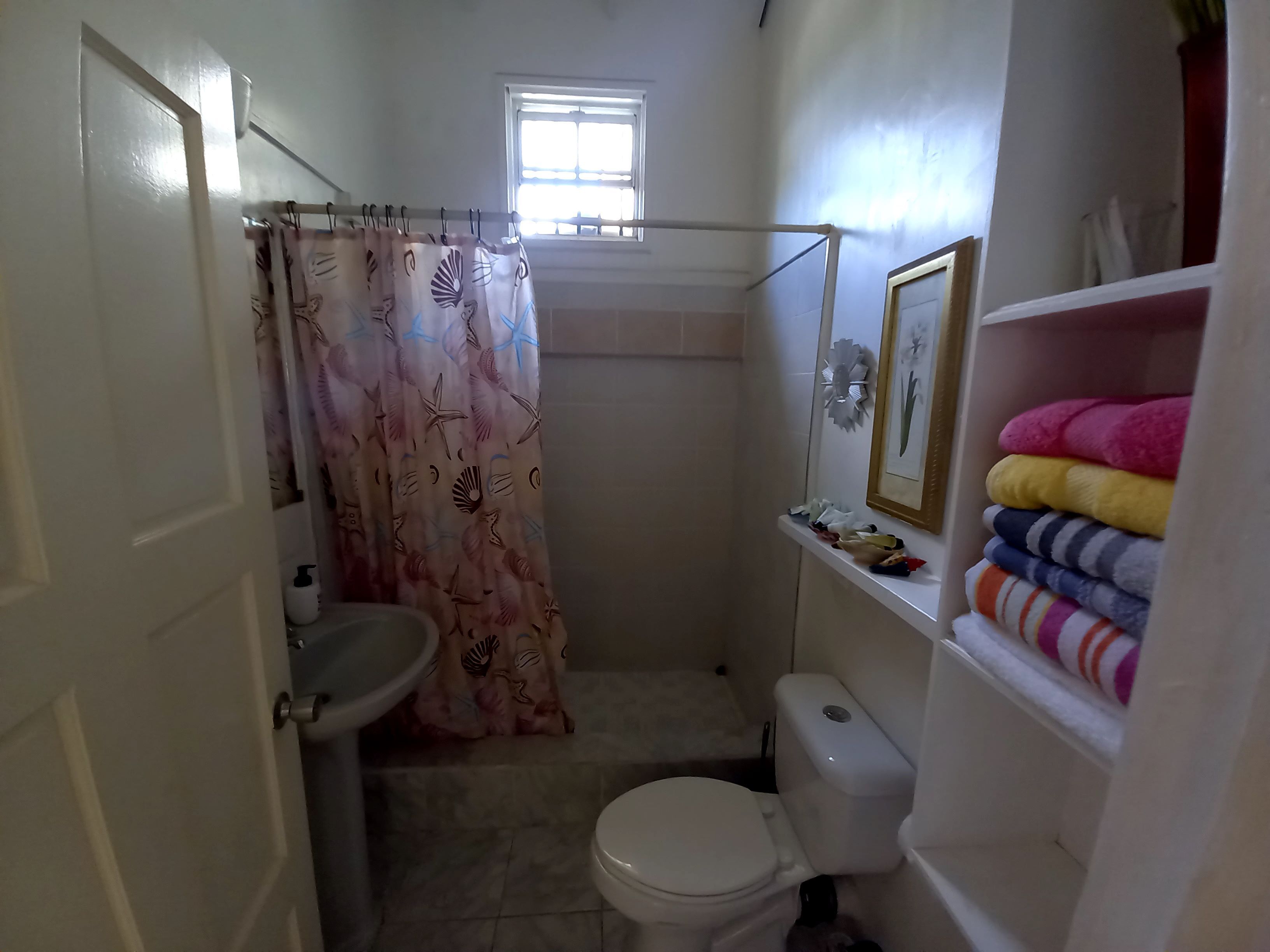 A small bathroom with a toilet, sink, and a shower curtain with a sea life pattern. There is a window above the shower, and a shelf with folded towels.