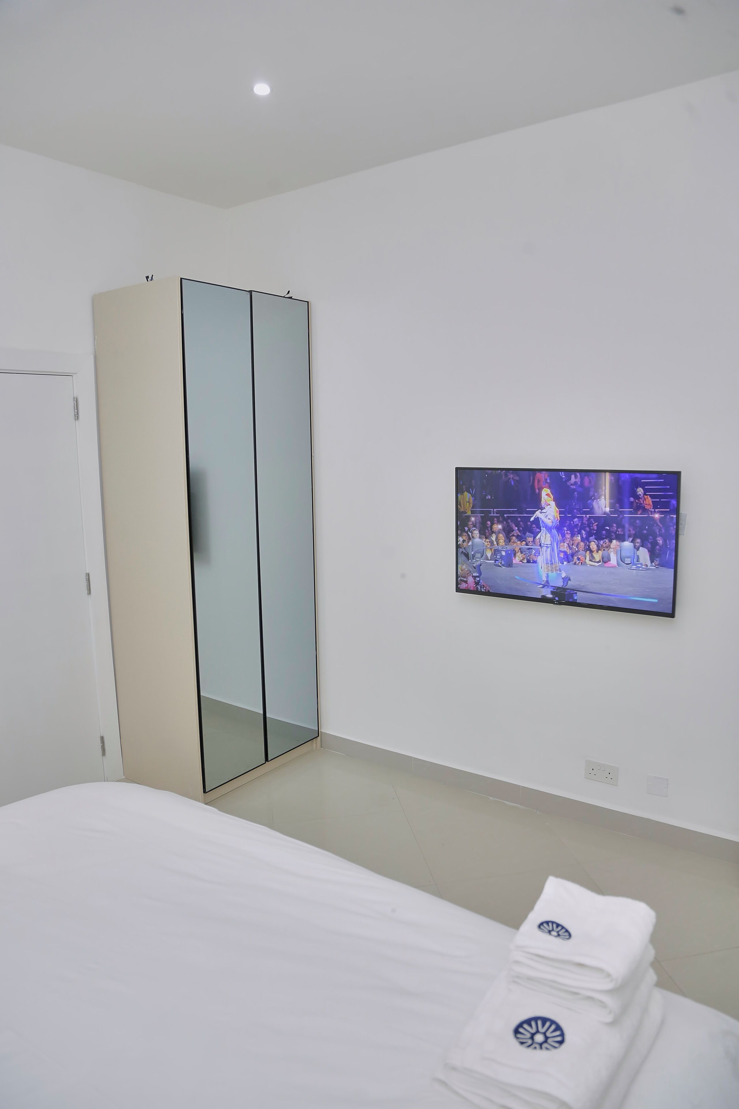 A hotel room with a bed, a TV mounted on the wall, a wardrobe, and a stack of towels.
