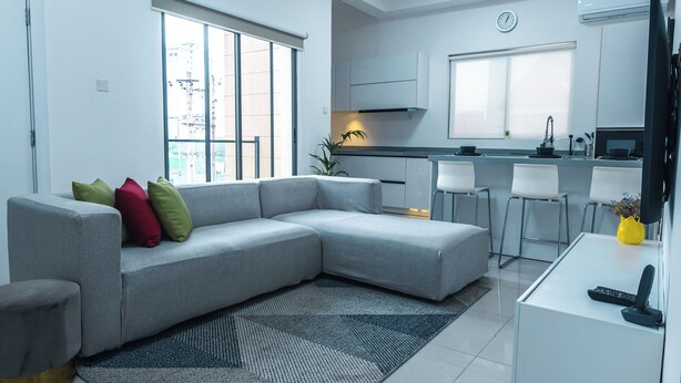 En moderne stue med en gr å hjørnesofa, en lille rund skammel og en sort-grå måtte. I baggrunden ses et køkken med hvide skabe samt et spiseområde med barstole.