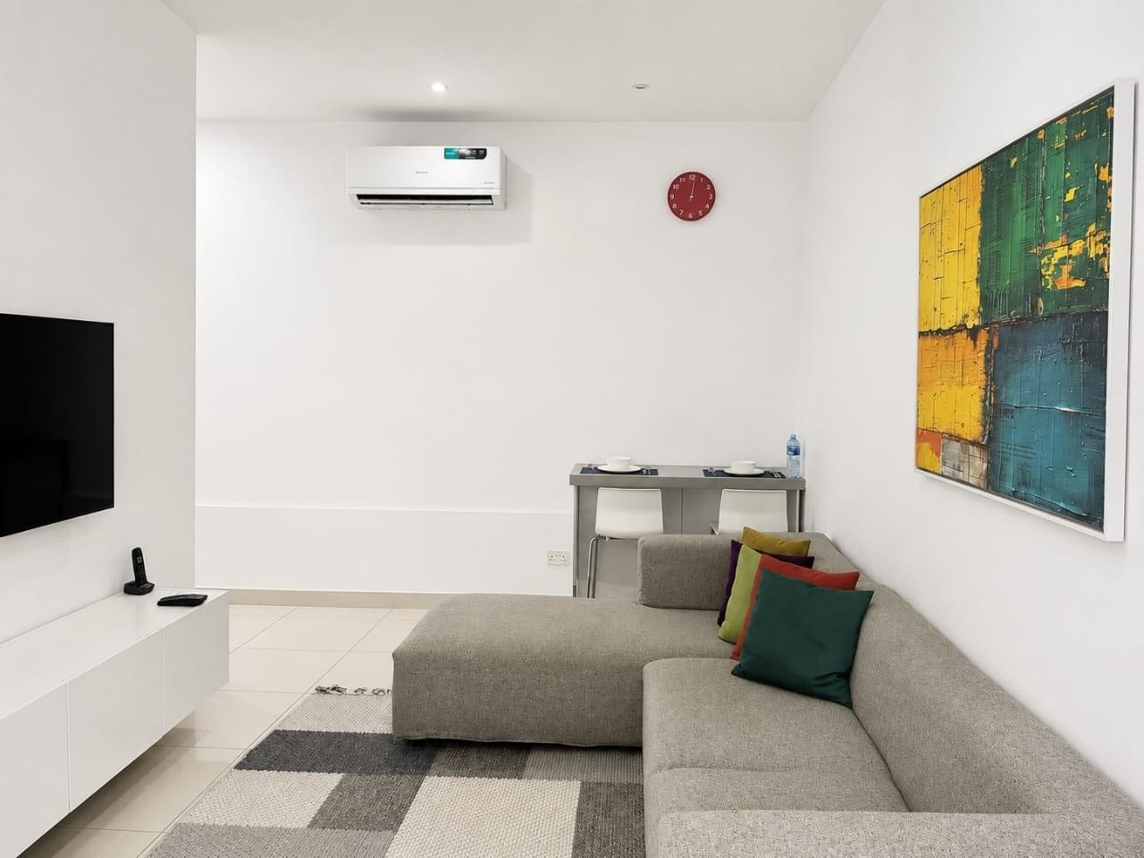 A modern living room with a flat-screen TV, a gray sofa, a dining table with chairs, and a colorful abstract painting on the wall.