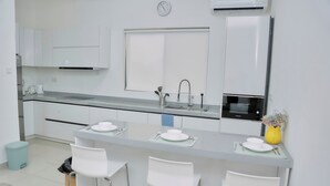Et moderne køkken med hvide skabe, en indbygget mikroovn og en vask. Der er tre hvide barstole og en lille skraldespand.