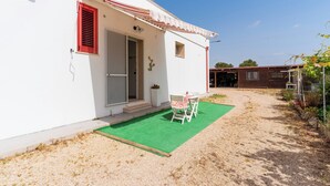 A white building with a red window shutter, a small porch with a table and chairs, and a green artificial turf area.
