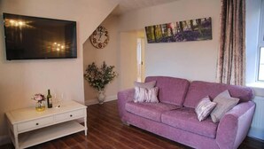 Un salón con un sofá morado, un mueble blanco para televisor, un reloj y un cuadro en la pared.