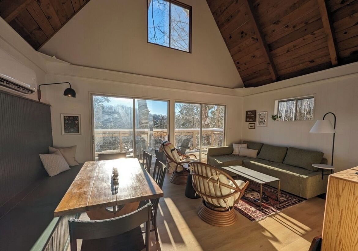 Een ruime woonkamer met een houten plafond, grote ramen met uitzicht op de omgeving, een eettafel met stoelen, een bank en een klein bijzettafeltje.