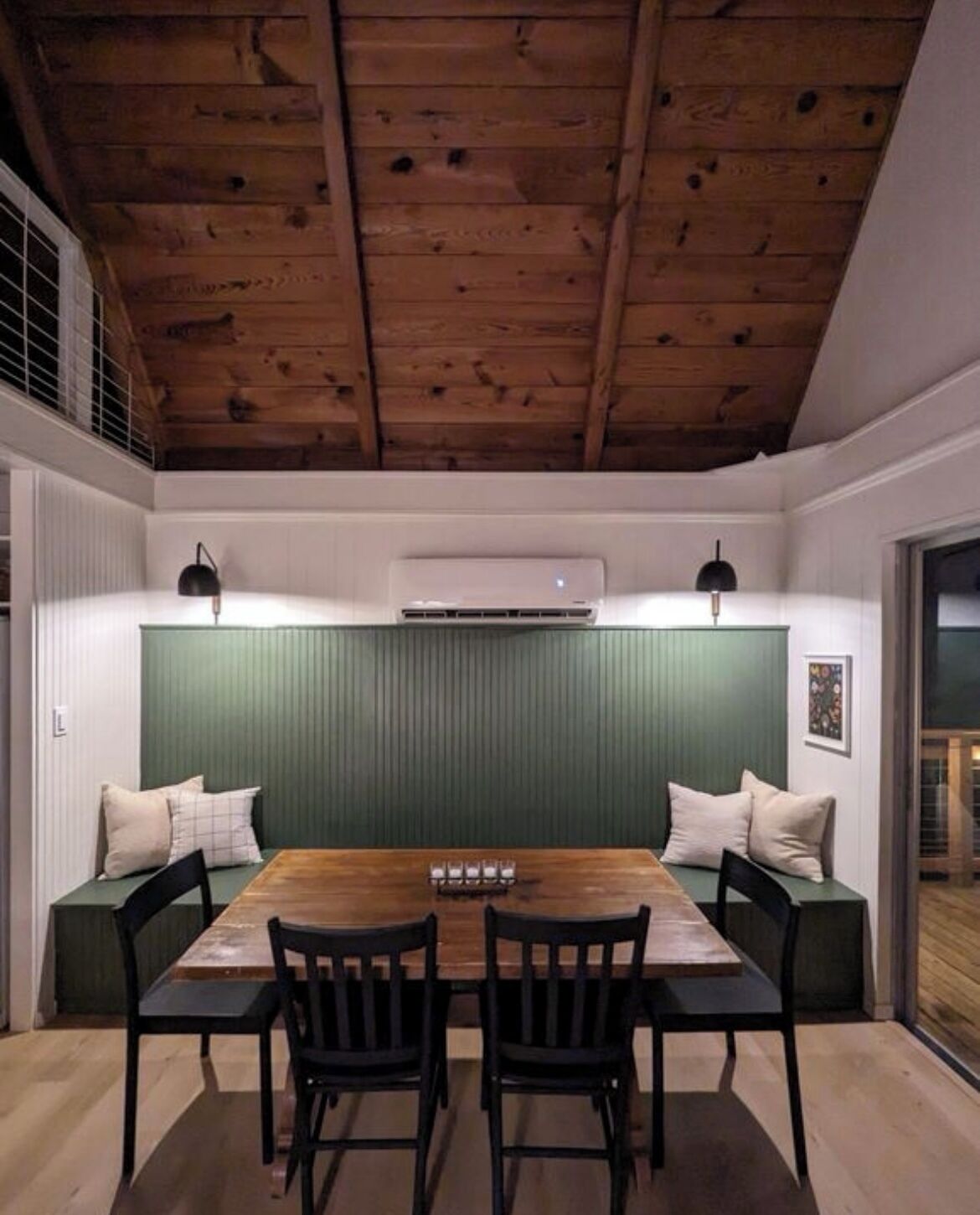 A dining area with a wooden table and chairs, a green upholstered bench, and a white ceiling with wooden beams.
