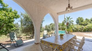 An outdoor patio with a table and chairs, surrounded by white columns, and overlooking a garden with trees and a pool. - Casamarmora Al Carrubo (Sicilia)
