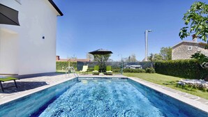 A swimming pool with a clear blue water surface, surrounded by a paved deck. There are lounge chairs and an umbrella providing shade.
