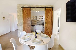 Un coin repas avec une table ronde dressée pour quatre personnes, des chaises blanches et une bouteille de vin.